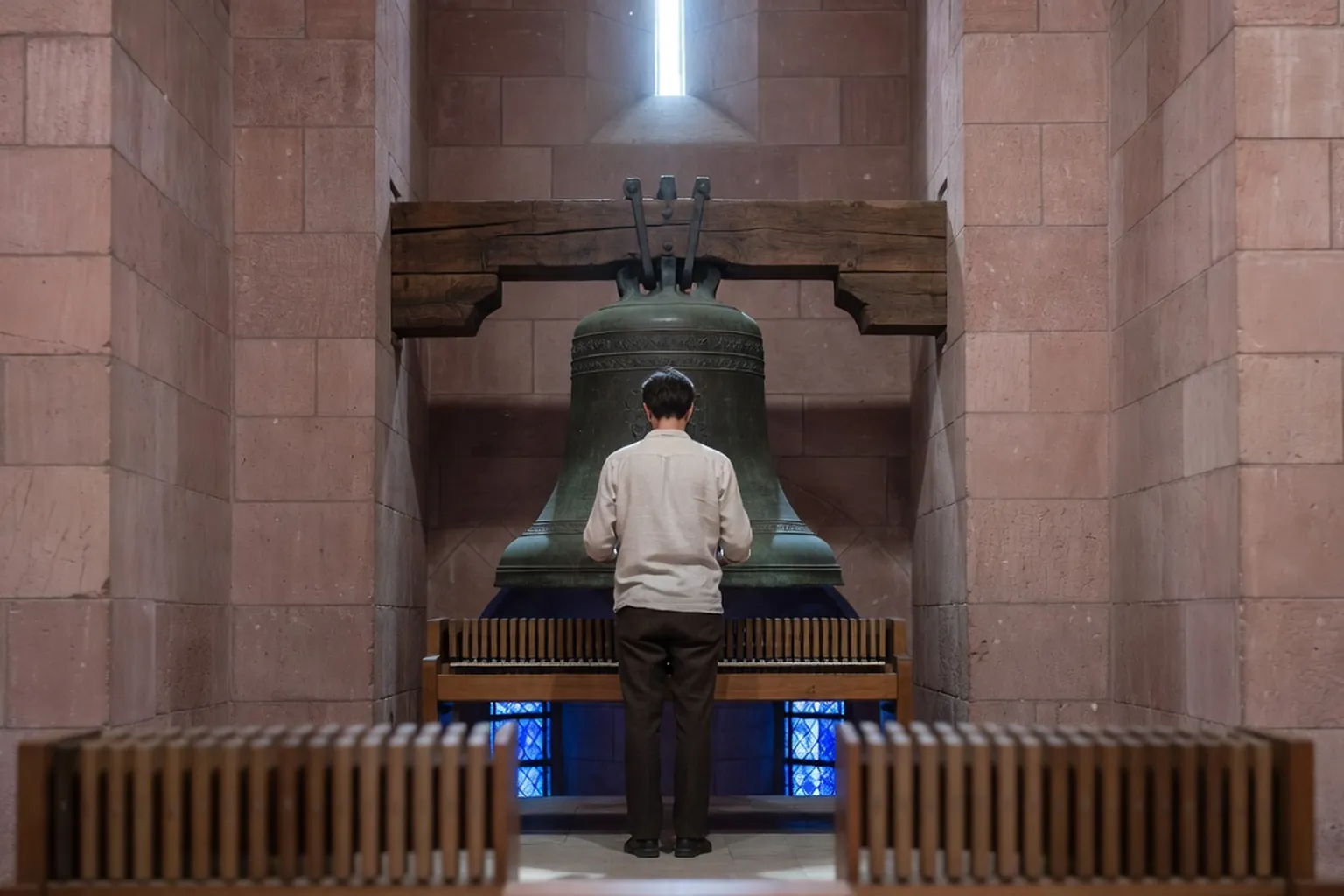 Carillonneur de dos debout face à une cloche de bronze ancienne suspendue dans un beffroi de grès rose, clavier à bâtons au premier plan